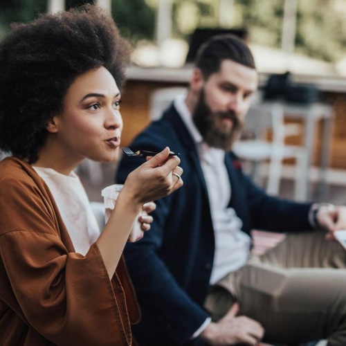 woman sits and eats outside while someone holding a cell talks to her
